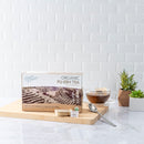 A package of Prince of Peace Organic Pu-Erh Tea, teabags on a wooden tray with a teaspoon and a cup of tea on a white countertop with some plants.