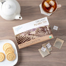 A package of Prince of Peace Organic Pu-Erh Tea, tea bags, a teapot, cookies on a plate, and tea in a cup with ice on a table.