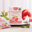 A box of Prince of Peace Peach Oolong whole leaf tea in front of a peach, next to styled on a light surface with loose tea sachets, a triangle tea bag, and leaves. 