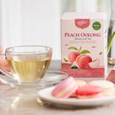A box of Prince of Peace Peach Oolong whole leaf tea stands next to a glass cup of brewed tea on a table, with pink macarons blurred in the foreground.