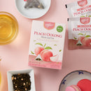 A box of Prince of Peace Peach Oolong Whole Leaf Tea is arranged on a pink surface with tea bags, brewed tea, loose tea leaves, and macarons around it. 