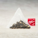 A triangular tea bag filled with loose-leaf tea sits on a light beige surface, with a small red tag showing the 'Prince of Peace' brand name beside it.