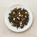 A top-down view of loose-leaf tea mixed with small dried peach pieces, presented on a round white ceramic plate against a textured light beige background.