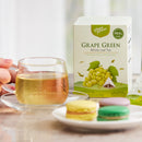 A box of Prince of Peace Green Grape Whole Leaf Tea with a cup of tea aside with  some macarons on a plate in the front.