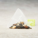 A single triangular tea bag filled with loose-leaf grape green sits on a light neutral surface, with a small green tea bag tag reading 'Prince of Peace' beside it.