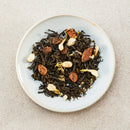 A top-down view of Prince o Peace Grape Green Tea on a light blue-gray ceramic plate, featuring green tea leaves mixed with dried green grape, gren tea leaves, and small beige botanical pieces, set against a neutral textured beige background.