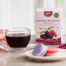 A box of Prince of Peace Mulberry Rose Black Whole Leaf Tea stands on a tabletop beside a glass cup of tea, with purple and pink macarons in the foreground.