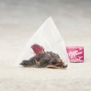 A triangular tea bag filled with dark dried berries, tea leaves, and a red flower, placed on a light beige surface next to a pink box labeled 'Prince of Peace'.