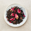 A top-down view of loose-leaf tea in a small white ceramic bowl, featuring dark tea leaves mixed with bright pink rose petals and dried mulberry pieces, set against a neutral beige textured background.