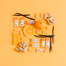 Packaging of Ginger Soft Chews arranged with oranges, ginger, and vanilla beans on a yellow background.