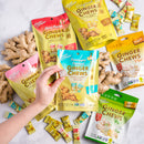 A hand holds a Prince of Peace Pineapple Coconut Ginger Chew in front of several Prince of Peace Ginger Chew pouches and scattered individual wrapped pieces on a light marble surface.