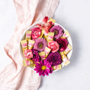 A white bowl filled with pink and purple flowers and Prince of Peace Ginger Chew Lychee individually wrapped pink and yellow candies, styled on a white surface with a light pink linen napkin.