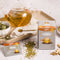 A package of Prince of Peace Immune Tea, a sachet, ingredients, dryer leaves around with a teapot, and a spoon on a tray.