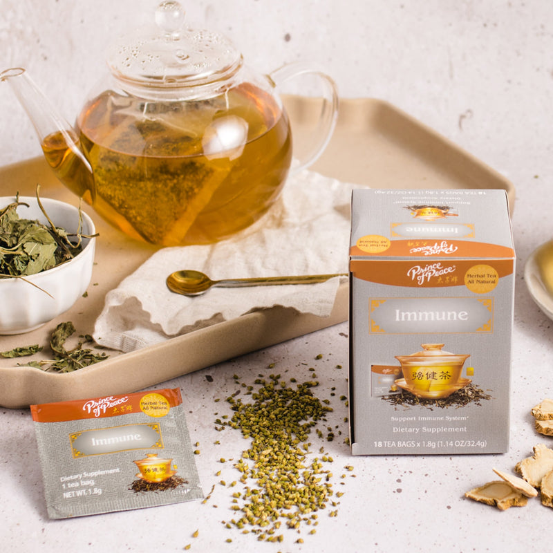 A package of Prince of Peace Immune Tea, a sachet, ingredients, dryer leaves around with a teapot, and a spoon on a tray.