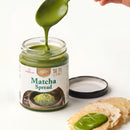 A jar of Matcha Spread with green matcha paste dripping from a spoon, with a few crackers topped with the spread in the foreground on a clean white background.