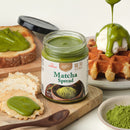 A jar of Matcha Spread is shown on a wooden board with matcha toast, waffles topped with green sauce and cream, a small bowl of loose matcha powder, and a wooden spoon, all on a clean white background.