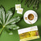 Prince of Peace Organic Green Tea in a cup with a package, tea bags and tree leaves aside on a green background.
