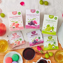 A flat-lay (top-down) product display featuring three Prince of Peace Fruit Whole Leaf Tea boxes with their corresponding individual foil sachet packets, arranged on a textured cloth and wooden tabletop alongside tea accompaniments.