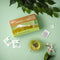 A glass of Prince of Peace Premium Japanese Style Green Tea with a package ,and tea bags and some green leaf on a green background.