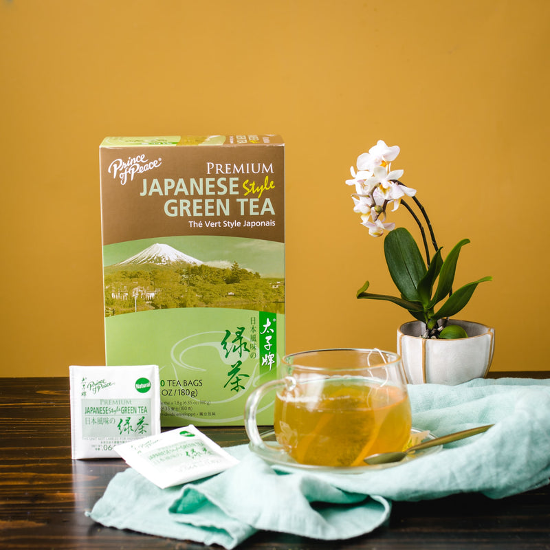 A glass of Prince of Peace Premium Japanese Style Green Tea with teabags, a package and an orchid on a table..
