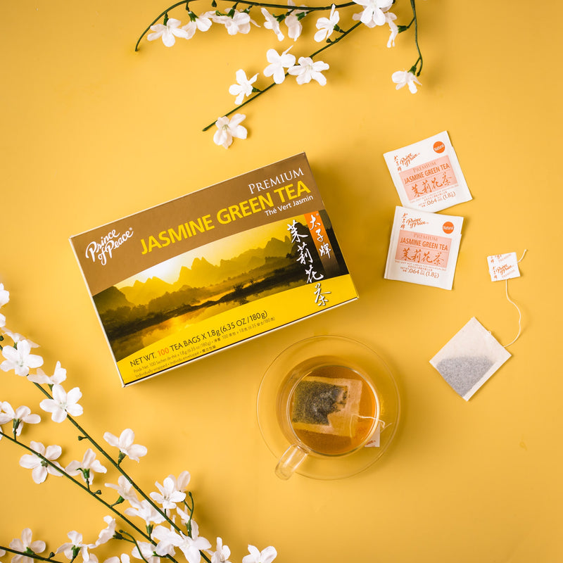 A glass of Prince of Peace Premium Jasmine Green Tea with some teabags, a package,and jasmin on a yellow background.