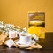 A cup of Prince of Peace Premium Jasmine Green Tea with a wooden tea spoon, tea bags in a bowl, jasmin and a package on a table.