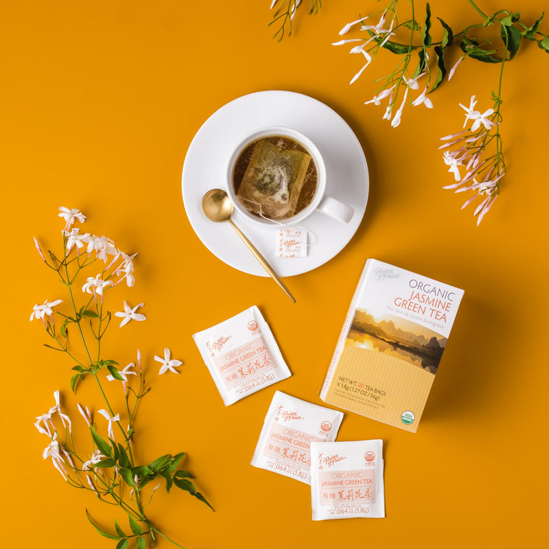 A cup of Prince of Peace Organic Jasmine Green Tea with a package and tea bags, jasmine aside on an orange background.