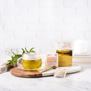 A glass of Prince of Peace Organic Jasmine Green Tea with a package and a tea bag, a spoon, jasmine aside on a wood tray.