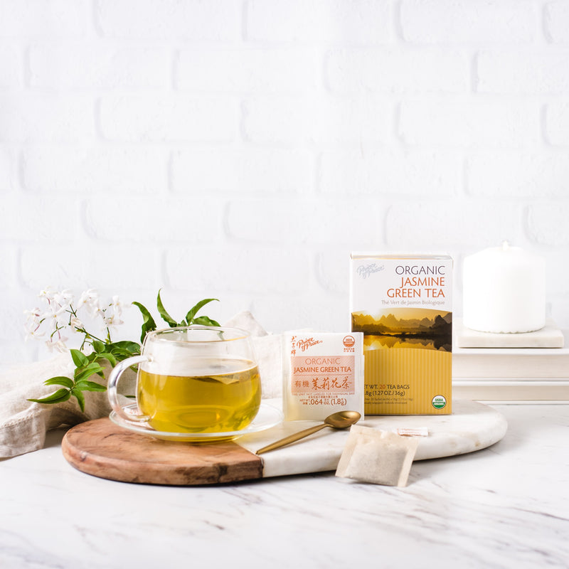 A glass of Prince of Peace Organic Jasmine Green Tea with a package and a tea bag, a spoon, jasmine aside on a wood tray.