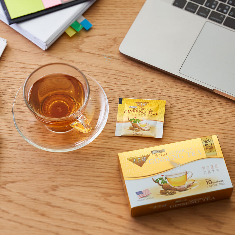 Prince of Peace Wild American Ginseng Instant Tea inner box, sachet and a cup of tea with a laptop and notebook on a desk..